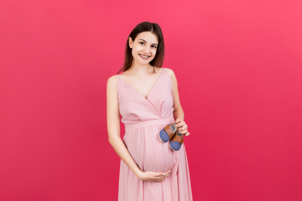 快乐的孕妇展示小靴子的一个小男孩在彩色背景。未来的母亲在等孩子。本空间图片下载