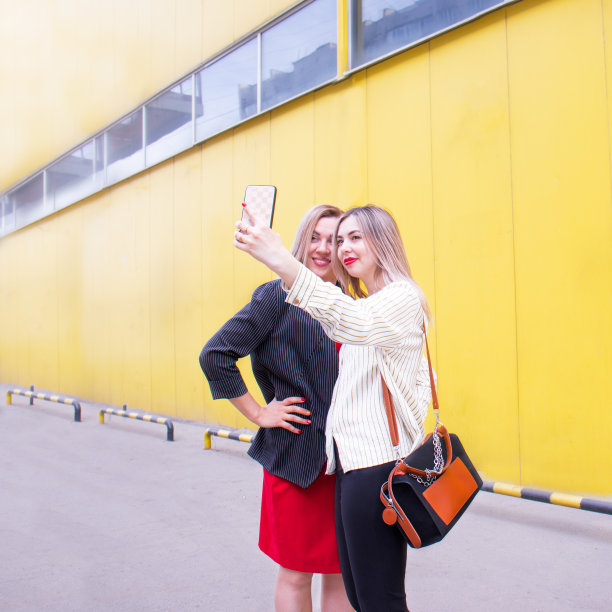 两个漂亮的年轻金发女人用智能手机对着一堵黄色的墙自拍。侧观，女性友情图片下载