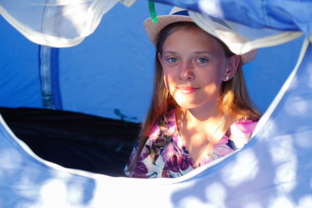 女孩舒适地坐在蓝色露营帐篷里，透过防蚊网窗向外看，戴帽少女的肖像，旅行儿童，阳光照射，放松休息，背景虚化图片下载