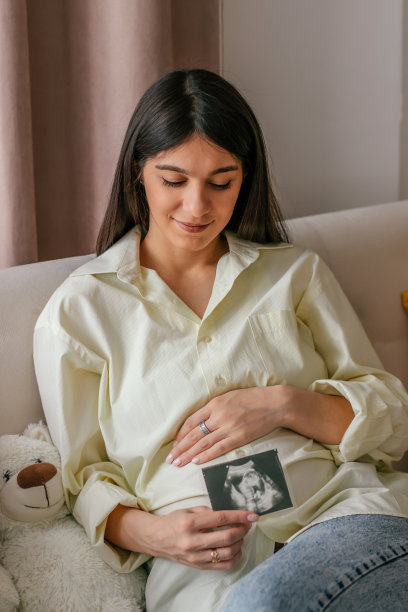 身穿黄色衬衫和牛仔裤的年轻孕妇坐在家里的沙发上，手里拿着一张超声波照片。妊娠及产前护理理念。图片下载