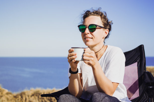快乐的年轻女子旅行家戴着太阳镜，拿着铁杯子和茶或咖啡，在自然景观中享受大海风景，坐在营地。旅行露营和冒险生活理念。本空间图片下载