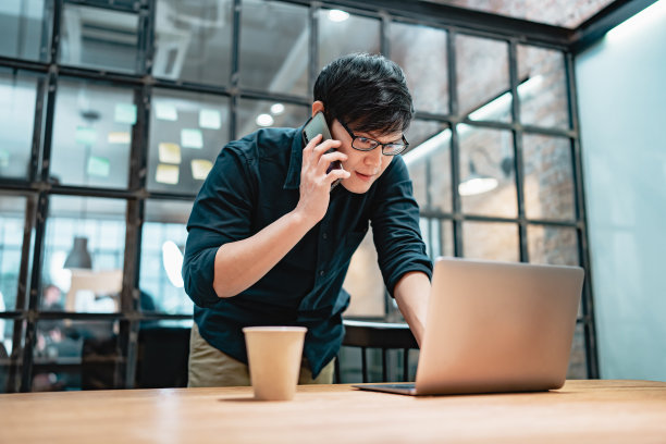 在现代办公场所，亚洲商人坐在笔记本电脑前用手机交谈。员工在工作时用智能手机聊天。图片下载