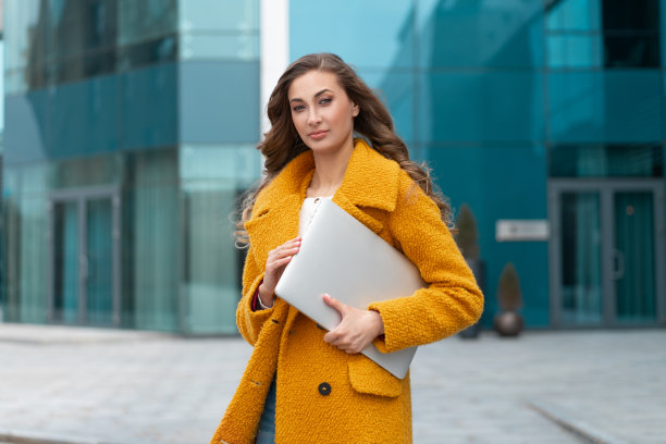 商务女性与笔记本电脑穿着黄色外套站在户外企业建筑背景图片下载
