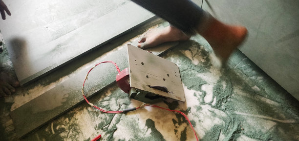 正在进行中的工作——一名木匠在建筑工地用圆形电锯工作， 与绿色的木板和木屑一起凌乱地散落在电线和机器附近，一个人光着脚行走图片下载