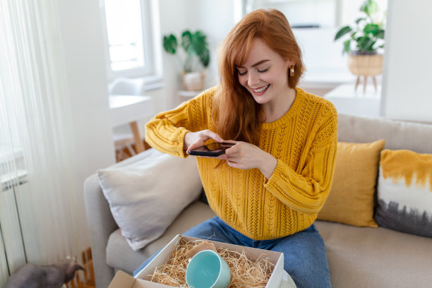 年轻满意快乐的女人购物狂客户坐在沙发上拆包裹送货箱，网上购物送货概念。为产品拍照，然后发布在社交媒体上图片下载