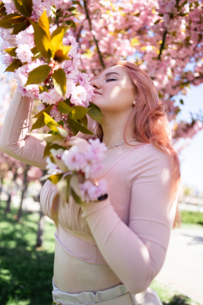 年轻美丽的时尚女孩在一个阳光明媚的日子里戴着帽子摆姿势附近盛开的树与粉红色的花的肖像。春天，女孩在一棵开花的樱花树旁。图片下载