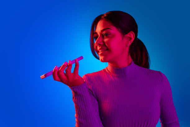 年轻快乐的女孩的肖像录音语音信息电话孤立的蓝色背景在粉红色的霓虹灯图片下载