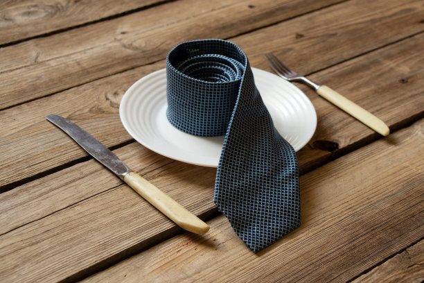一条蓝色领带放在一张木桌上的盘子上，旁边放着一把叉子和一把刀，一顿商务午餐，旁边是一条男士领带图片下载