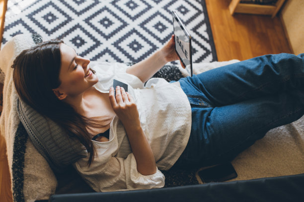 年轻女子坐在沙发上，用她的平板电脑和信用卡网购。图片下载