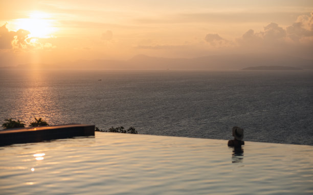一个女人的后视图与帽子放松在无限游泳池，看着美丽的日落和海景图片下载