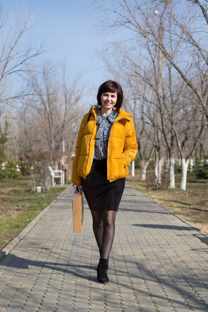 一位身穿黄色夹克的40岁至45岁的快乐女性拿着一台新笔记本电脑走在街上，这是一个购物概念图片下载