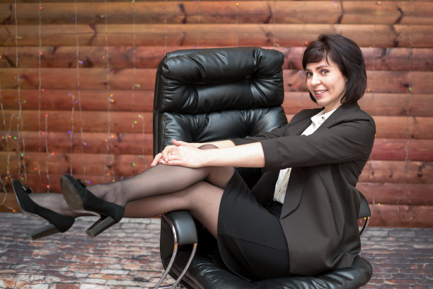 办公室里，一位面带微笑的神秘黑发女性坐在靠木墙的大皮椅上图片下载