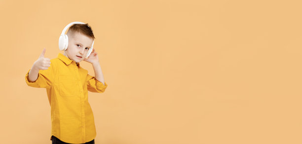 横幅的肖像有趣聪明的学校男孩戴着耳机在黄色衬衫。黄色工作室背景。教育。面带微笑，对着镜头竖起大拇指。高质量的照片图片下载