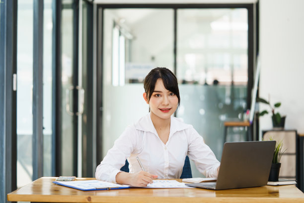 在现代办公室的办公桌前，穿着休闲的亚洲年轻女性微笑着看着镜头。图片下载