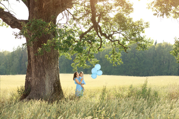 在一棵巨大的老橡树下，一位妇女带着一个带着蓝色气球的孩子。在一个喜庆的阳光明媚的夏日，妈妈和女儿一起在大自然中散步图片下载