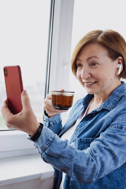 快乐的老年妇女在智能手机上进行在线视频通话，使用应用程序进行交流，站在窗边喝茶。与远在国外的子孙交谈。竖版图片下载
