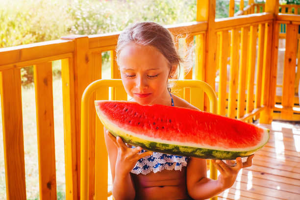 夏日露台上的女孩笑着吃了一大块红色的西瓜。自然的食物。女孩在夏天吃一个成熟多汁的西瓜。可爱的金发女孩在户外吃一片西瓜。图片下载