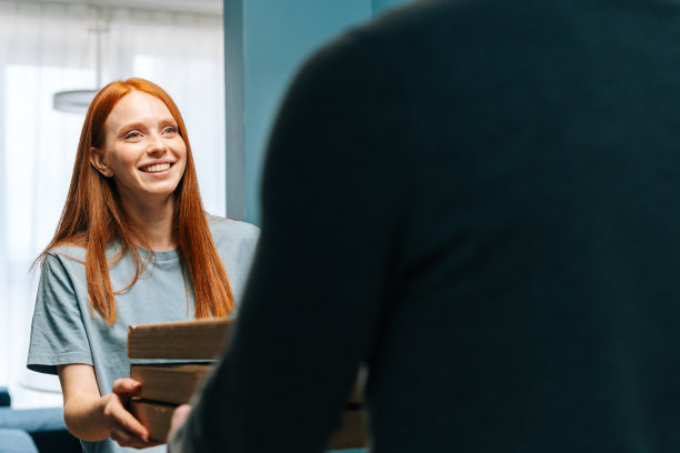 微笑的年轻女子收到纸盒与热披萨从不知名的送货员在家门口家门口。图片下载