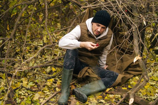 一个饥饿的徒步旅行者，穿着雨衣坐在森林树下的灌木丛中，手里拿着野生的红浆果，在阴雨的日子里看着它们。图片下载