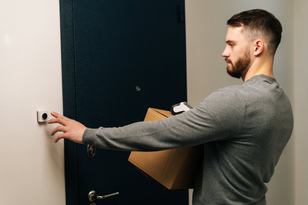 送货员手持纸板箱和非接触式支付POS无线终端按客户公寓门铃的背影。图片下载