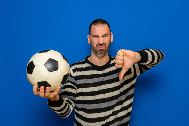 英俊的拉丁男子拿着足球与愤怒的脸，消极的标志显示厌恶与拇指向下，拒绝概念。孤立在蓝色背景。图片下载
