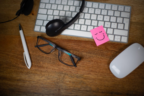 工作中幸福和幸福的象征。配备电脑、眼镜和耳机麦克风的工作站。键盘上贴着一张带微笑的粉色便签。图片下载