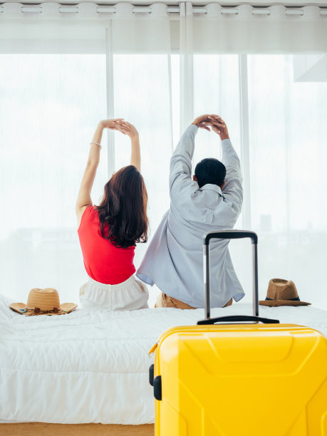 一对游客，节日快乐。暑假。年轻的亚洲男人和女人在床上伸展放松，手提箱靠近窗帘在酒店房间的窗户，垂直风格。图片下载
