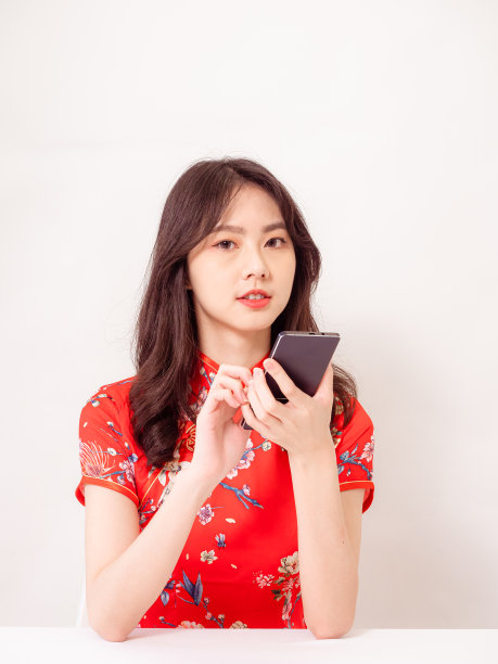 微笑的年轻亚洲女子在传统旗袍旗袍礼服使用智能手机孤立在白色背景。图片下载