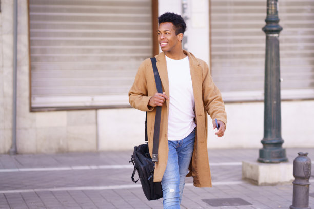 年轻的黑人男子走在街上，手里拿着公文包和智能手机。图片下载