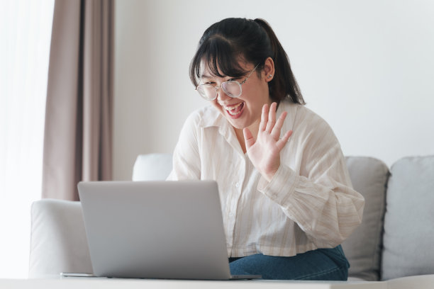 年轻的亚洲女性正在使用笔记本电脑进行在线视频会议电话，在客厅的沙发上挥手打招呼。图片下载