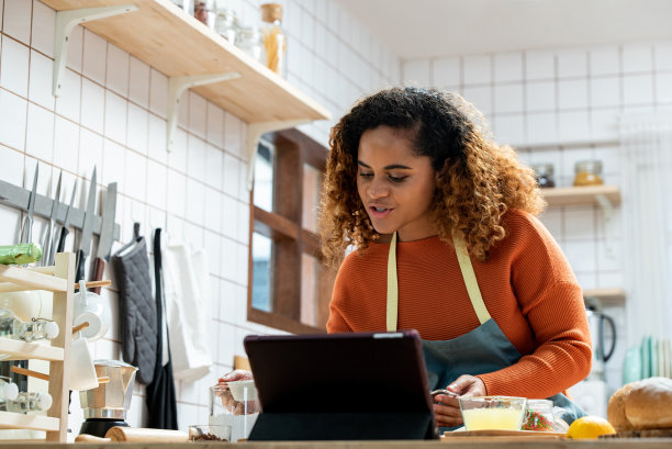 身穿围裙的非裔美国女性视频博主站在桌子旁，低头看，同时创建在线食品准备课程图片下载