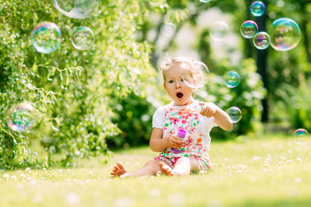 一个小女孩坐在公园的草地上吹肥皂泡。快乐童年的概念图片下载