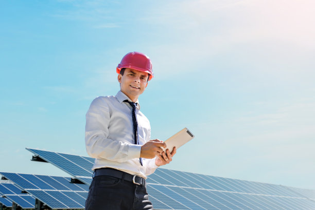 年轻的工程师身穿白色衬衫，头戴红色安全帽，手持平板电脑，正在验证太阳能发电厂的光伏板。图片下载