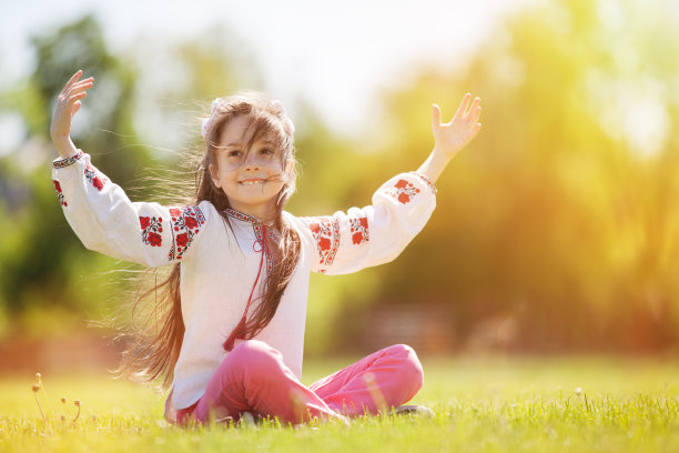 可爱的女孩在绿色的草地上穿着乌克兰民族服装微笑。画像中的快乐女孩伸出双手，坐在绿色的草地上。素材图片
