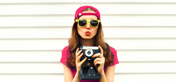 肖像快乐的年轻女子摄影师拍摄的电影相机和吹她的嘴唇戴着棒球帽的白色背景图片下载