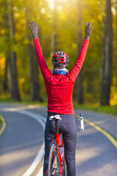 公路自行车赛的想法。迷人的女自行车手在温暖的服装摆在公路自行车，而转身与举起手在森林户外对秋天的道路背景。图片下载