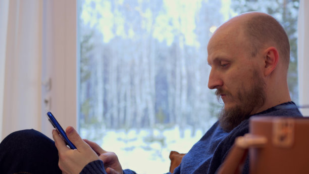 一个留着胡子的严肃男人坐在沙发上，聚精会神地看着他的智能手机，和别人聊天，不想一个人呆着，沉思地听着留声机里的古典音乐。图片下载