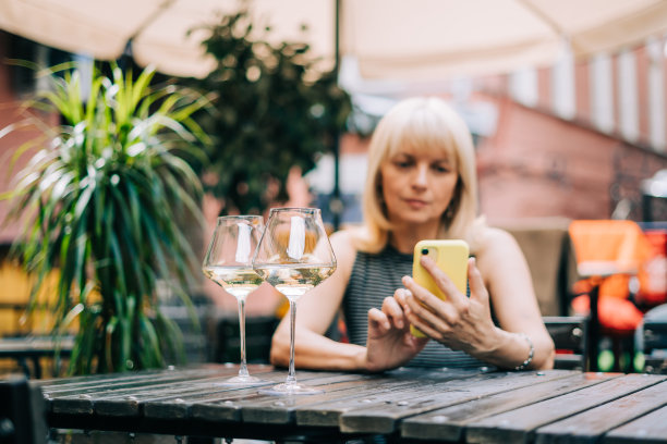 成年成熟的女人在酒吧户外用手机发短信，带着酒杯和模糊的餐厅，喝着白葡萄酒。使用科技手机的人。生活在夏日阳光明媚的日子图片下载