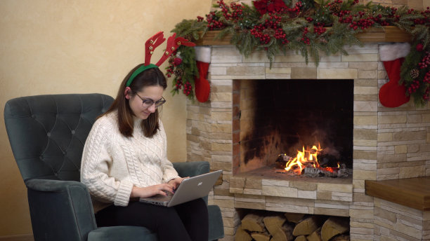 一位戴眼镜的年轻孕妇坐在壁炉旁的扶手椅上，头上顶着角，正在笔记本电脑上打字。女孩给圣诞老人写了一封电子邮件。图片下载