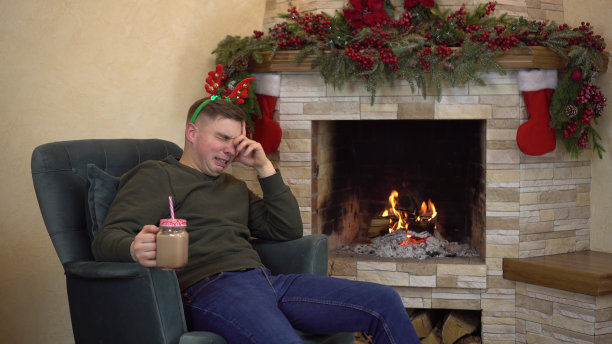 一个悲伤的年轻人坐在壁炉旁的扶手椅上，头上戴着犄角，一边喝着可可，一边哭泣。悲伤的圣诞节。图片下载