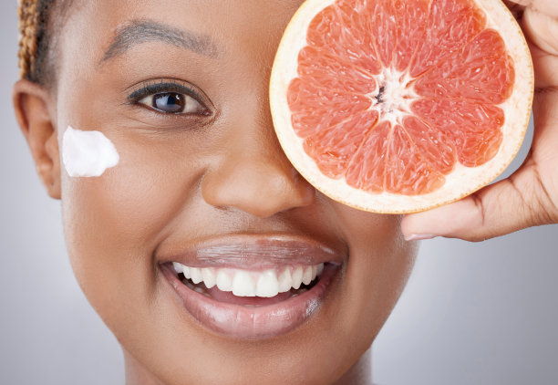 一位迷人的年轻女性拿着一片葡萄柚并涂抹保湿霜的影棚照片，背景为灰色图片下载