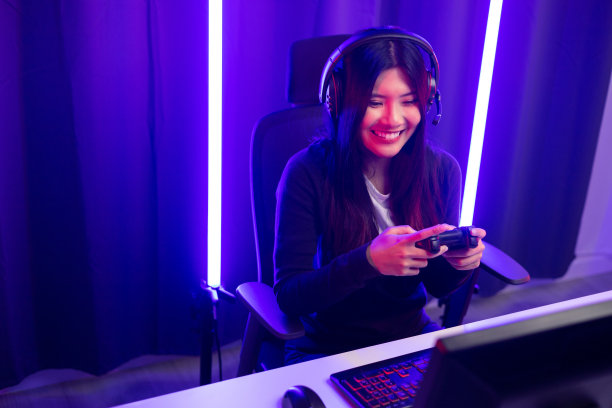玩视频游戏。年轻的亚洲漂亮女人坐在椅子上握着操纵杆在客厅。快乐女专业流光华人戴着耳机在黑暗的房间里玩霓虹灯游戏。图片下载
