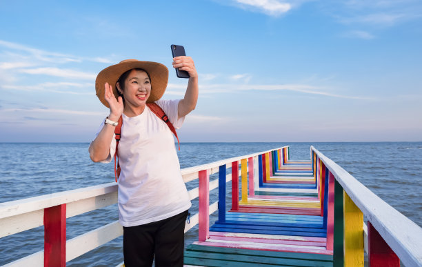 快乐的亚洲女游客挥手，而视频电话与智能手机在彩色木桥在海上的观点图片下载