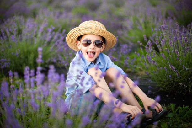 带着太阳镜微笑的小男孩坐在薰衣草地里。滑稽的孩子张开嘴巴吐着舌头，穿着一件蓝色的衬衫，戴着一顶草帽坐在薰衣草花之间。水平的照片图片下载