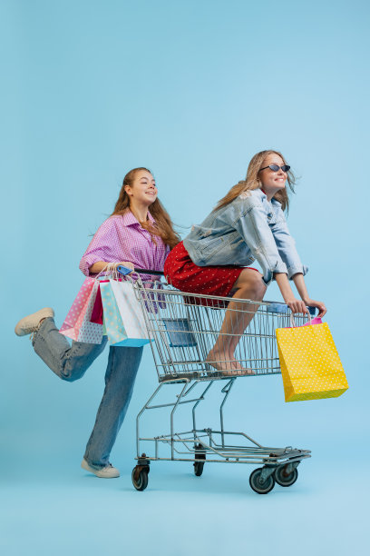 工作室拍摄的漂亮快乐的女孩与购物袋骑在一辆车孤立的蓝色背景。销售概念，黑色星期五，折扣，情感，青春和时尚图片下载