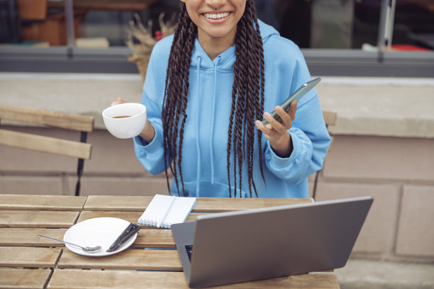 幸福的女性手里拿着一杯咖啡和手机，在自助餐厅的桌子上放着笔记本电脑。图片下载