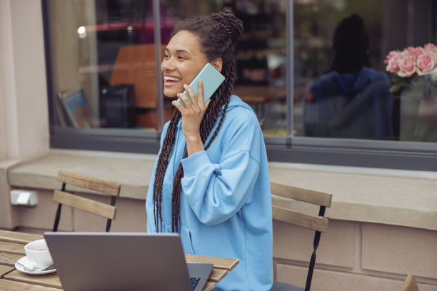 快乐的年轻混血女性在桌上的笔记本电脑上用手机交谈。咖啡馆露台。图片下载
