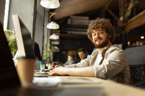高级开发人员。快乐的男人看着镜头，微笑着。在现代阁楼风格的办公室工作，在工作过程中使用设备和小工具。图片下载