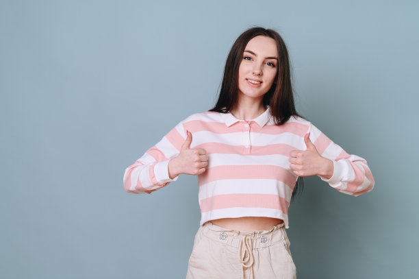 一个穿着条纹毛衣的活泼顽皮的小女孩在朴素的墙上竖起大拇指图片下载