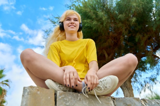 十几岁的女孩戴着耳机坐在公园的户外，蓝天白云图片下载